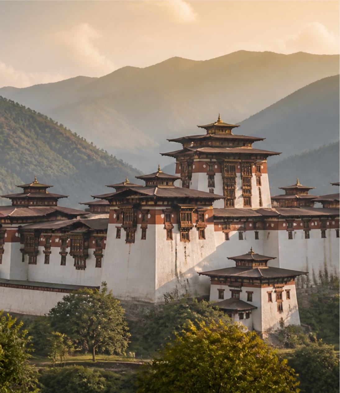 A Bhutanese dzong below forested Himalayan ridges in early light.